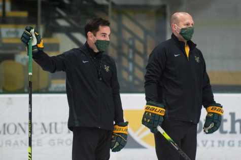 Head Coach Todd Woodcroft stands with Assistant Coach Stephen Wielder on the UVM ice rink holding hockey sticks.