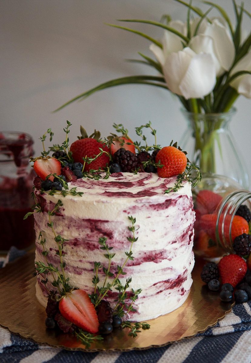 A vanilla chantilly cake filled with a mixed berry compote, frosted with whipped cream, and decorated with fresh berries and greenery. 